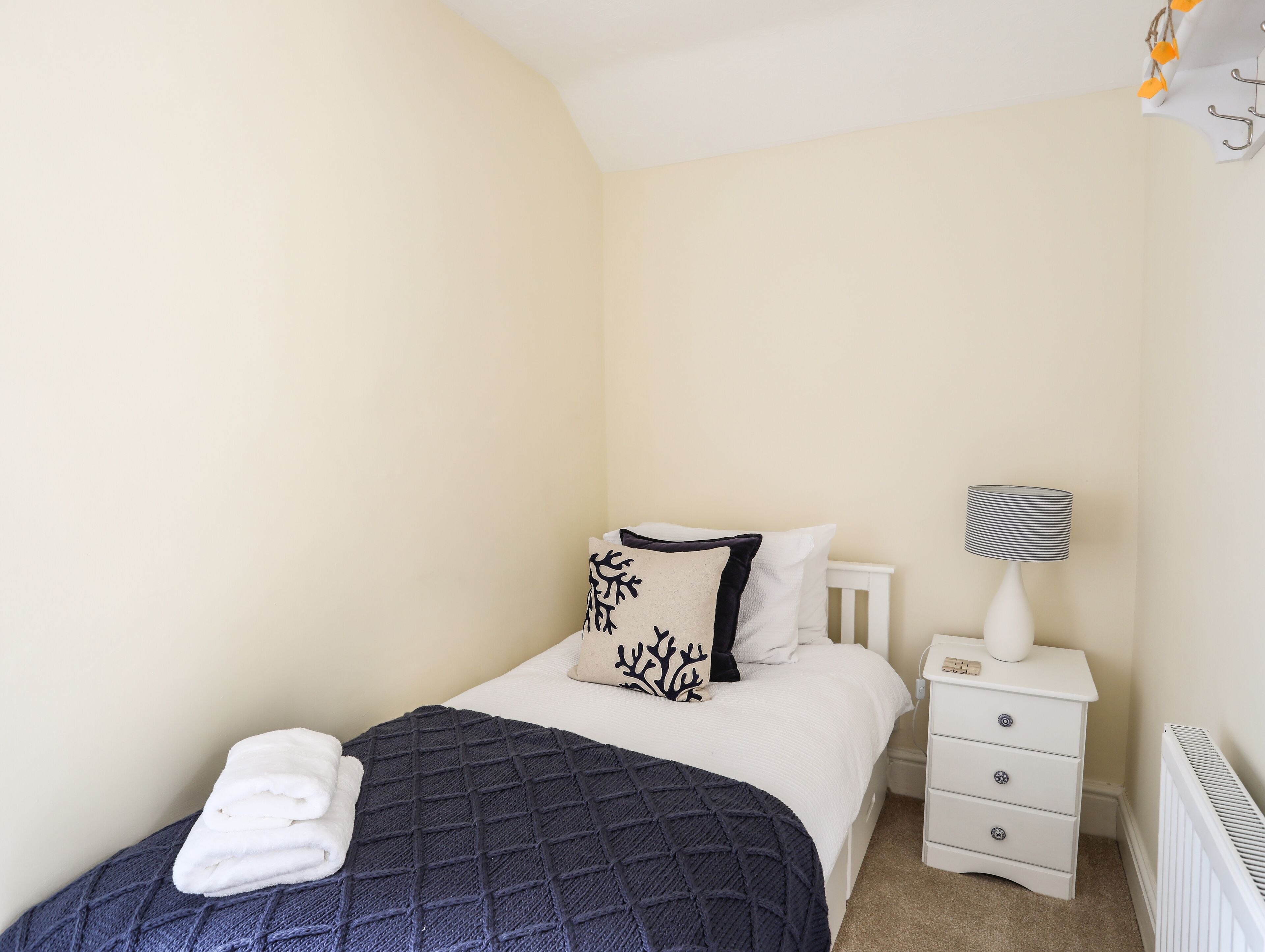 Een netjes opgemaakt bed met een donkerblauwe sprei, een wit kussen met een zwart boommotief, en een witte nachtkastje met een lamp.