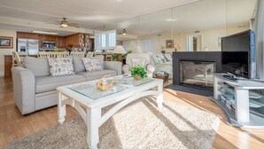 A spacious living room with a sofa, coffee table, and fireplace. A kitchen with wooden cabinets and a refrigerator is visible in the background. - Direct Oceanfront With a Pool! (Ocean City)