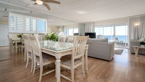 A spacious living area with a glass dining table, white chairs, a ceiling fan, a sofa, and a TV. - Direct Oceanfront With a Pool! (Ocean City)