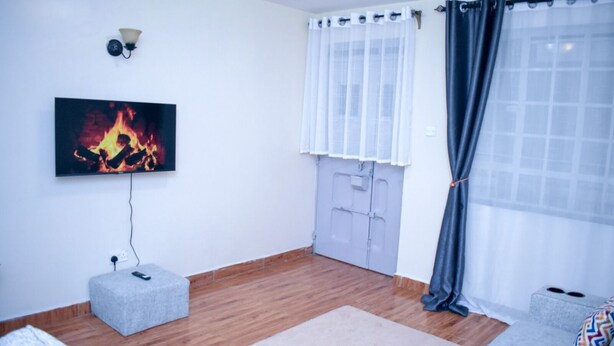 A living room with a flat-screen TV mounted on the wall, a grey ottoman, a wooden floor, a grey sofa, and a window with white curtains. - Dream house (Nairobi)