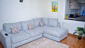 A grey sectional sofa with patterned cushions, a small wall-mounted light, a painting, and a kitchen area with cabinets and a microwave. - Dream house (Nairobi)