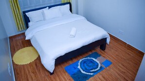 A neatly made bed with white linens and a rolled towel, a blue and white rug, and yellow curtains. - Dream house (Nairobi)