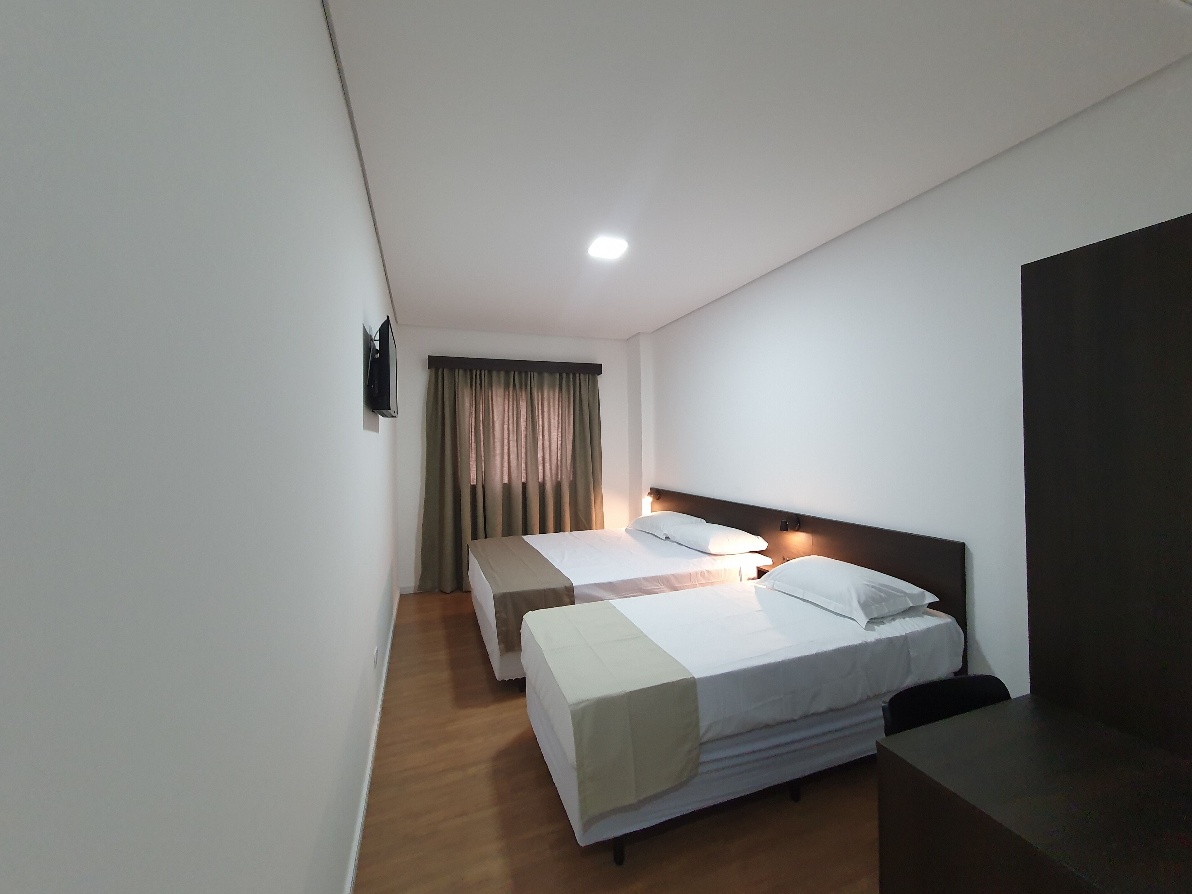 A hotel room with two single beds, a desk, a chair, a TV mounted on the wall, and a window with curtains.