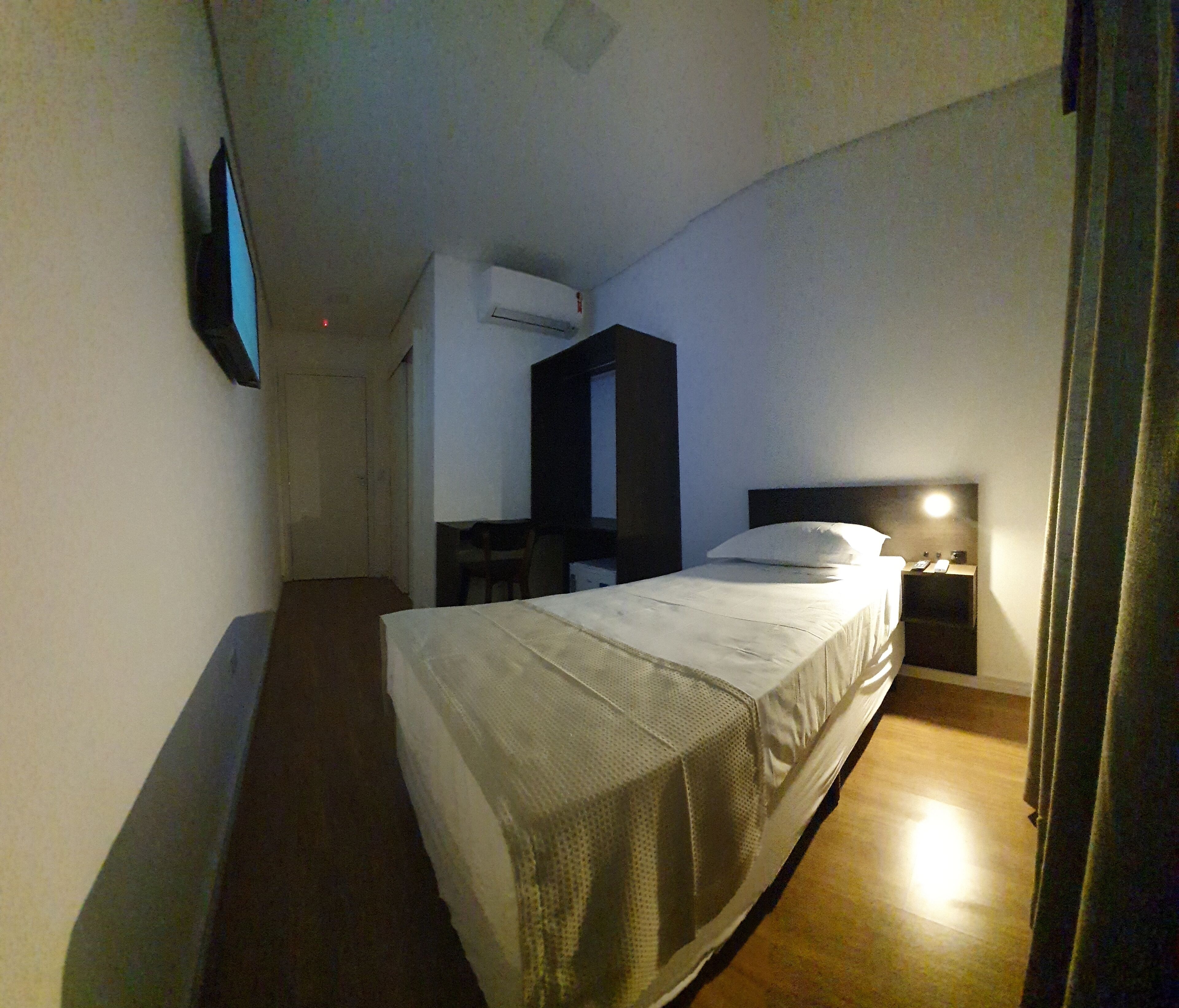 A narrow hotel room with a single bed, a desk, a chair, and a nightstand with a lamp.