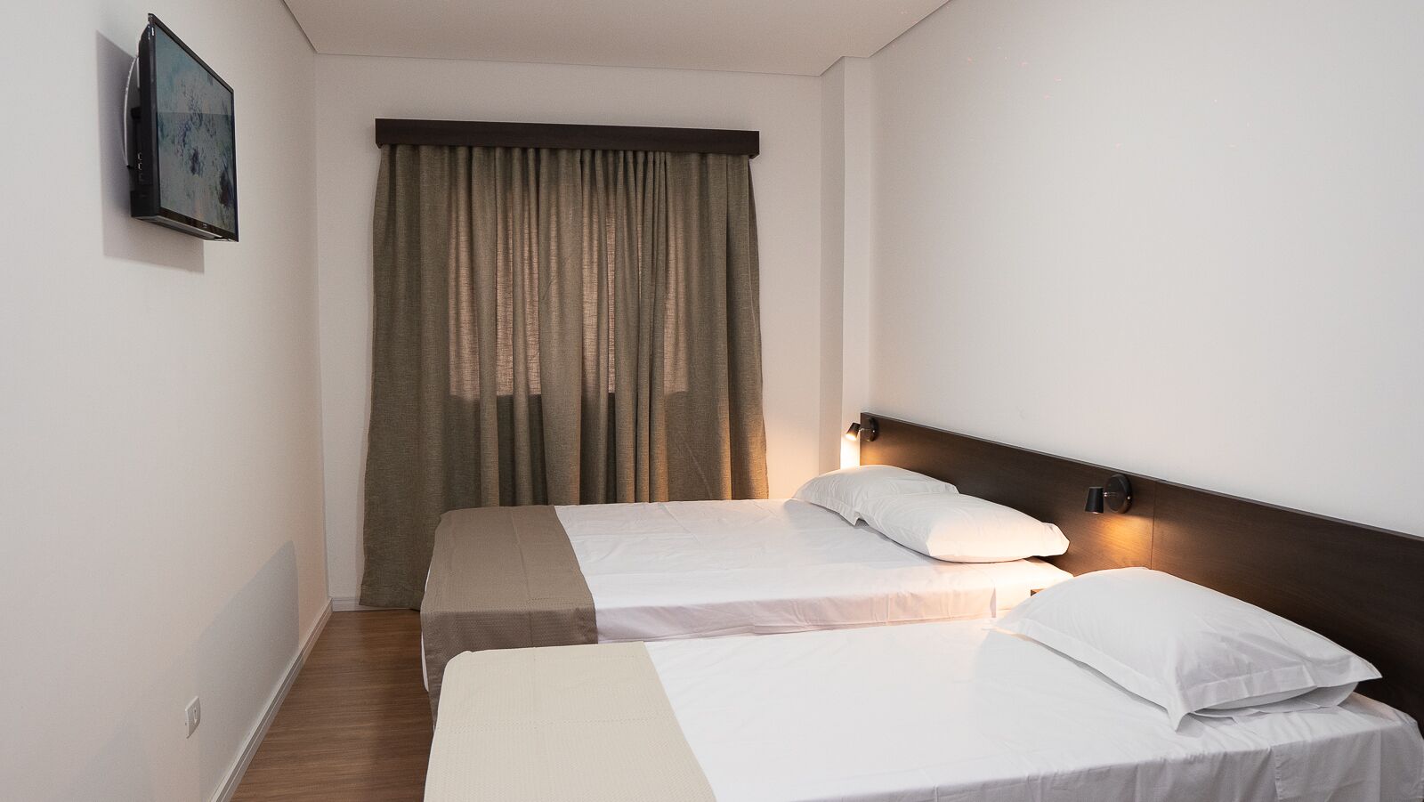 A hotel room with two beds, a large window with curtains, a mounted TV, and a wall-mounted light fixture.
