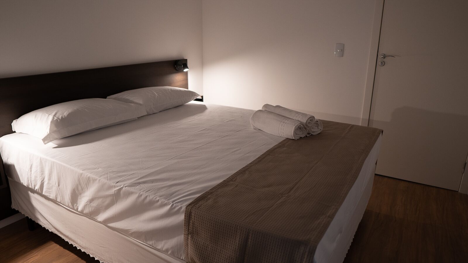 Cama bem arrumada com lençóis brancos e manta marrom, abajur e porta fechada em quarto de hotel.