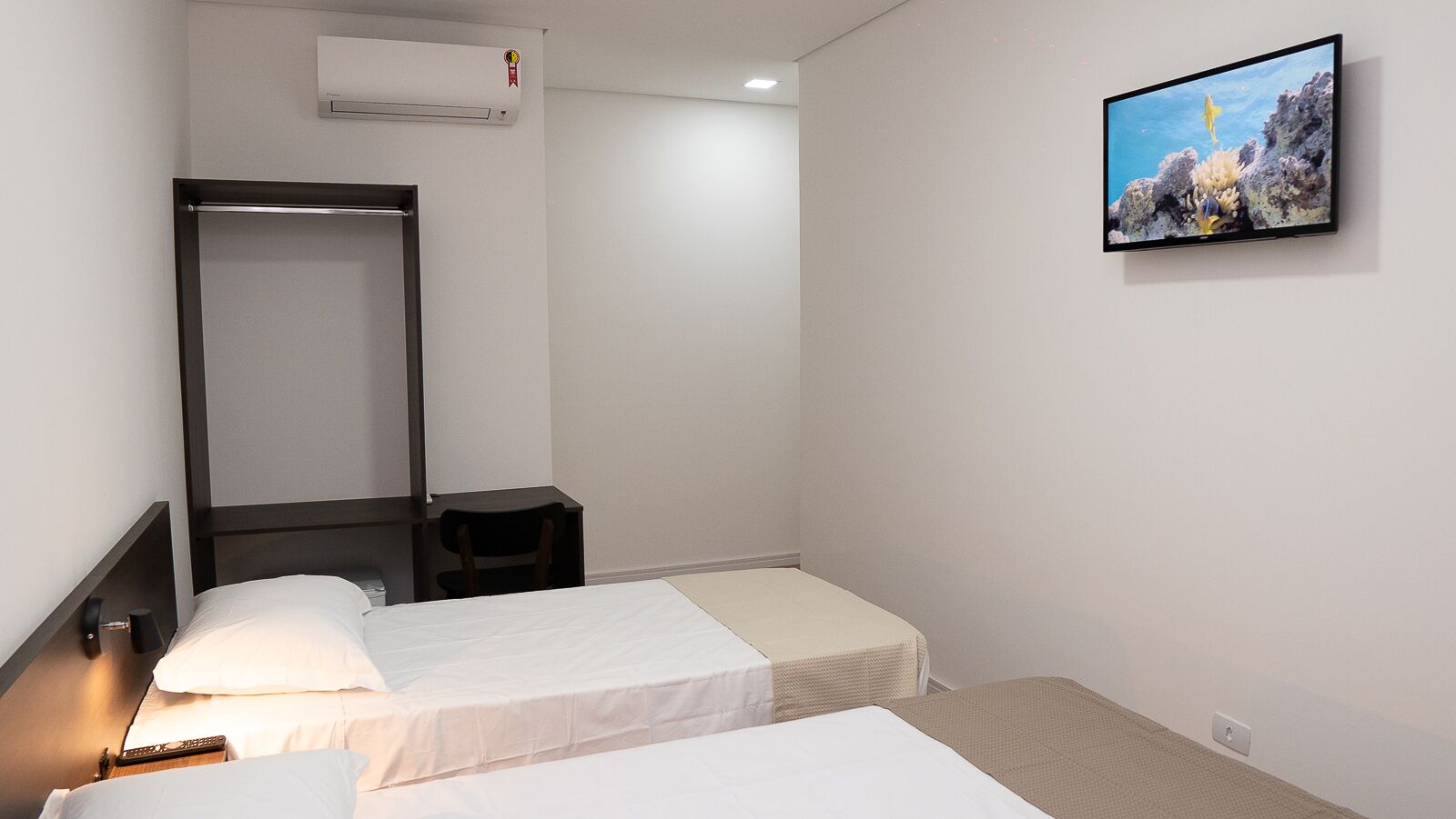 A hotel room with two single beds, a desk, a chair, a TV mounted on the wall, and a window with a view of a rocky coastline.