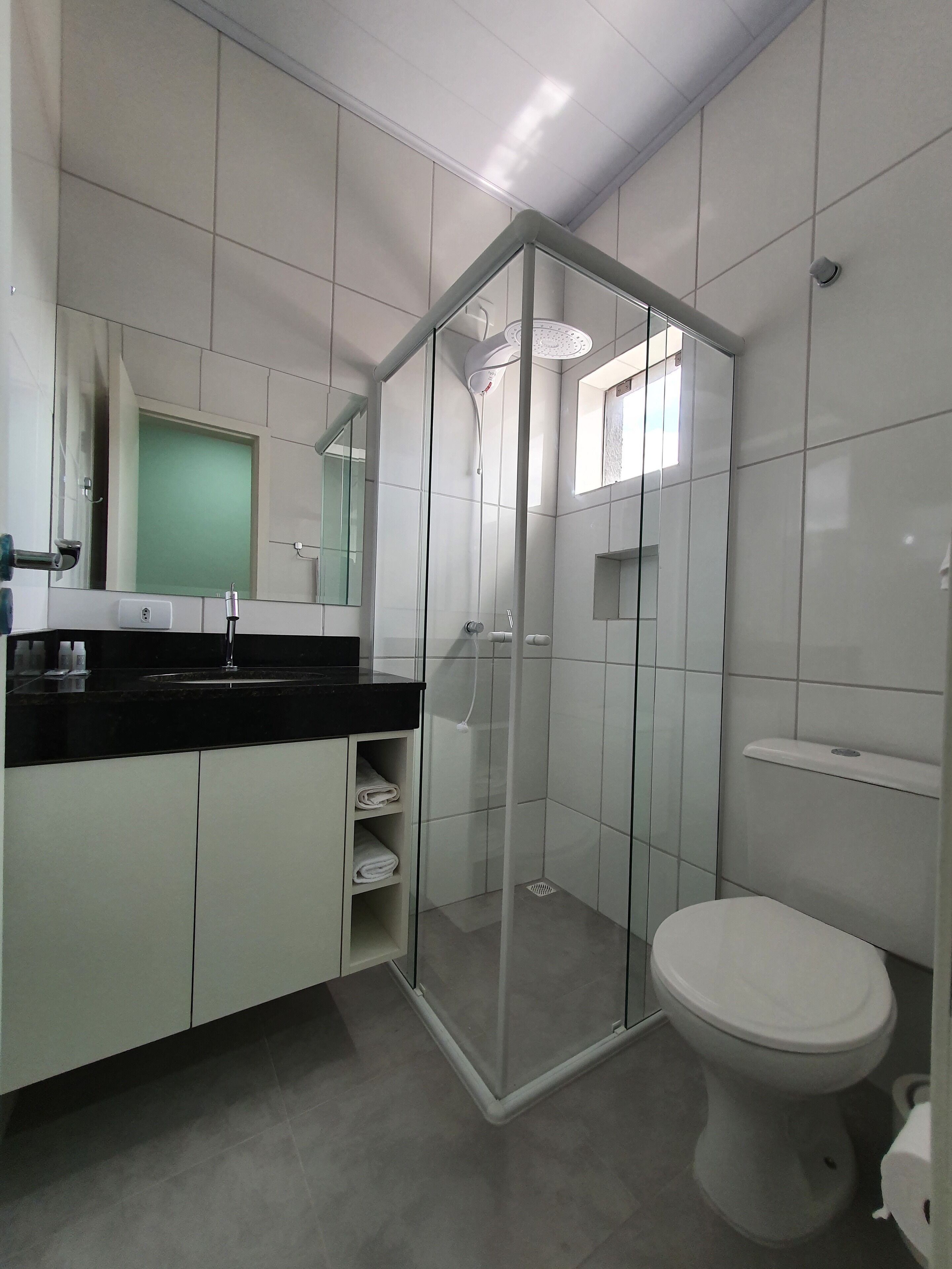 Une salle de bain moderne avec une douche à parois de verre, des toilettes blanches et un plan de toilette noir agrémenté d’un miroir et d’un porte-serviettes.