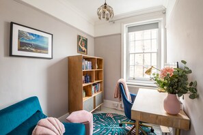 Une pièce chaleureuse avec un canapé bleu, une bibliothèque en bois, une table ornée d’un vase et une fenêtre donnant sur un beau paysage.