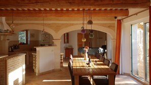 A spacious dining area with a wooden table and chairs, a kitchen in the background, and a large window providing natural light.