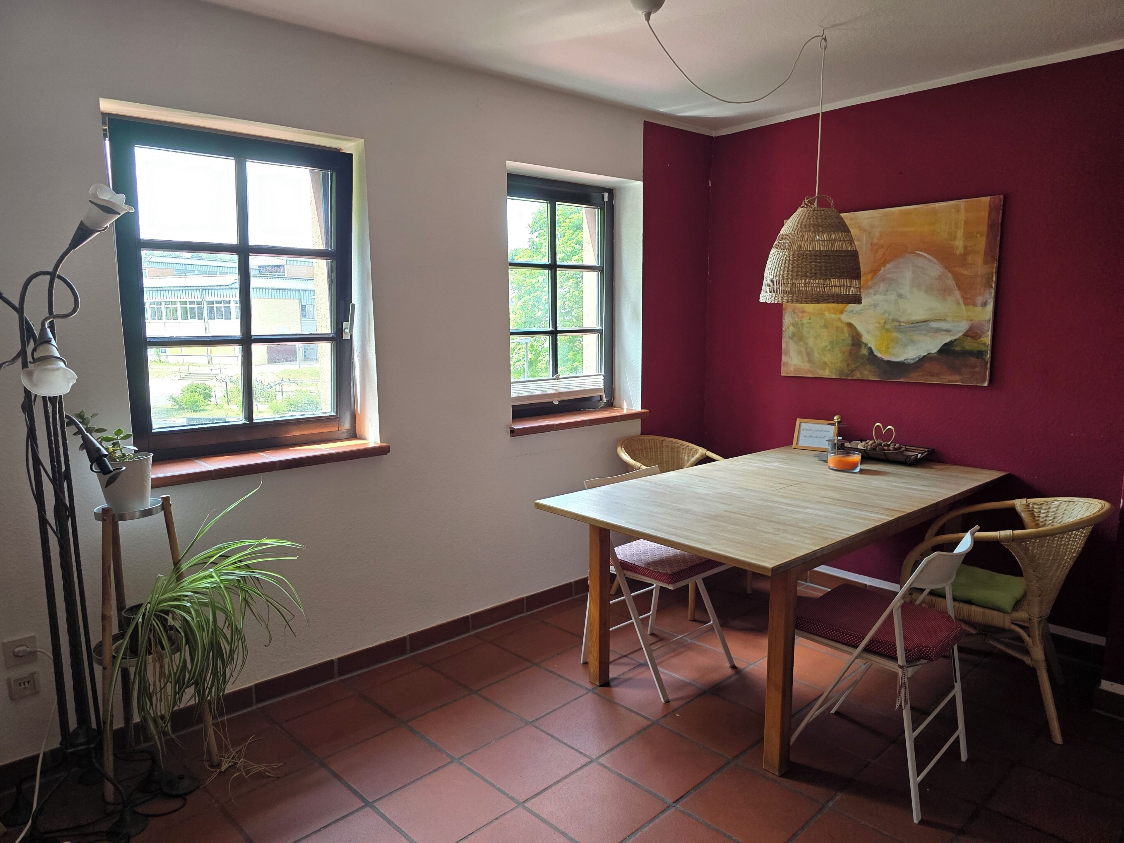 Un coin repas comprenant une table et des chaises, deux fenêtres, un luminaire suspendu et un tableau accroché au mur.