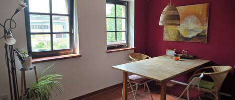 Un coin repas comprenant une table et des chaises, deux fenêtres, un luminaire suspendu et un tableau accroché au mur.