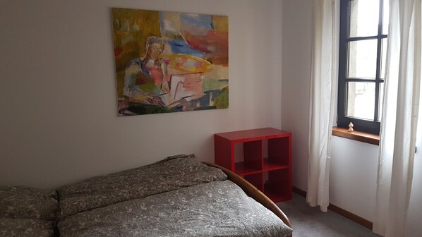 Una habitación con una cama, un estante rojo, una ventana con cortinas y un cuadro abstracto y colorido en la pared.