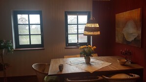 Un coin repas avec une table en bois, deux fenêtres, une lampe et un vase rempli de fleurs.