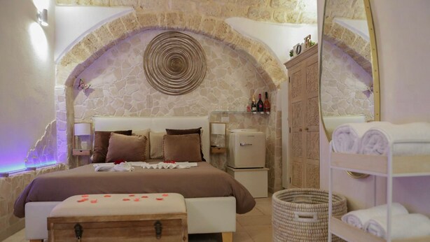 Une chambre confortable avec une arche en pierre, un lit parsemé de pétales de rose, un petit réfrigérateur et une commode en bois.