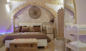 A cozy bedroom with a stone arch, a bed with rose petals, a small fridge, and a wooden chest.