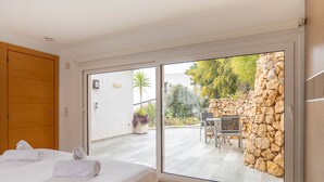 Et soveværelse med en seng, to sammenrullede håndklæder, en trædør og en skydedør af glas, der fører ud til en udendørs terrasse med en stenvæg, et bord og stole.