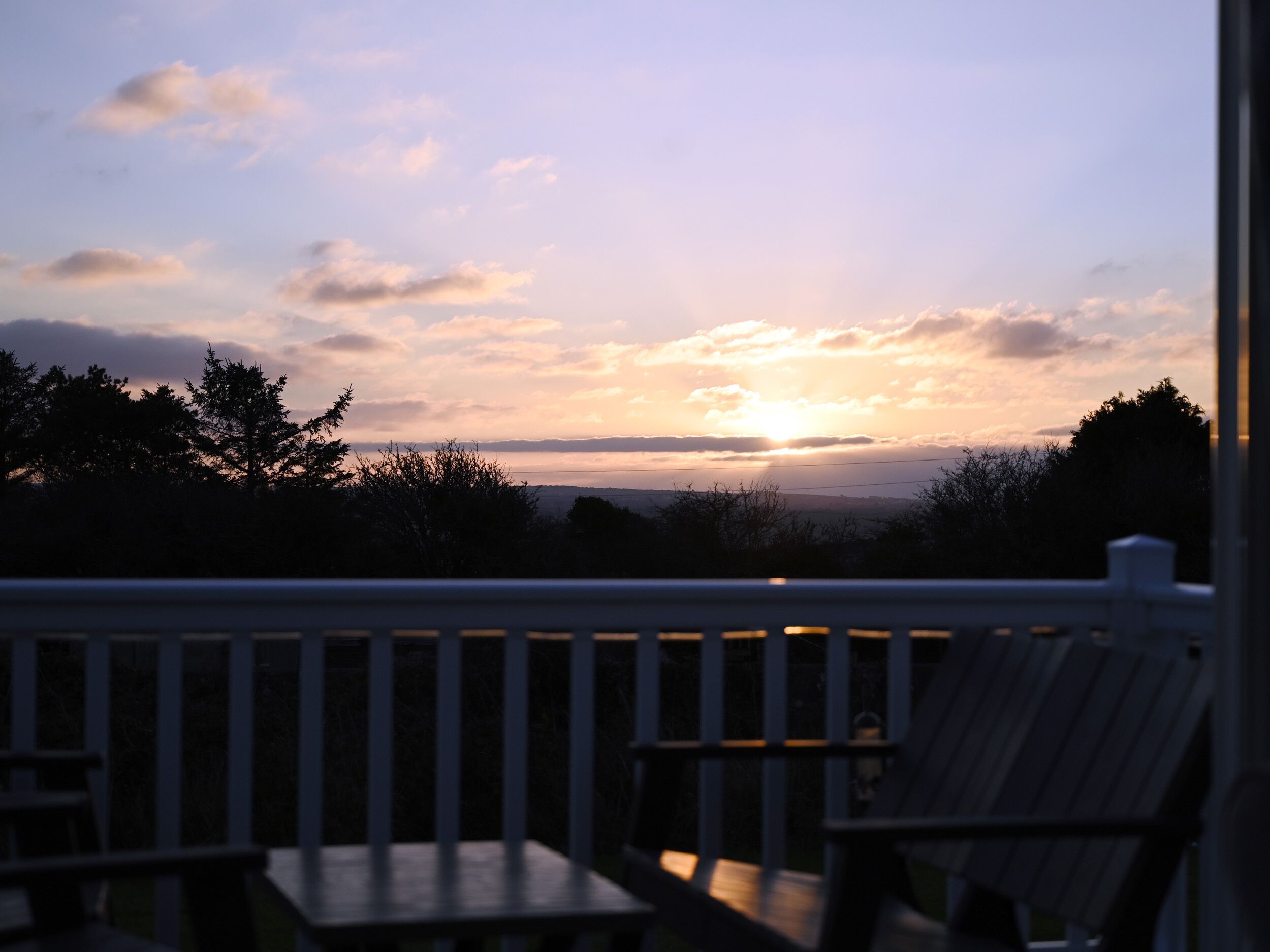 A deck with a table and chairs, overlooking a sunset over a distant landscape.
