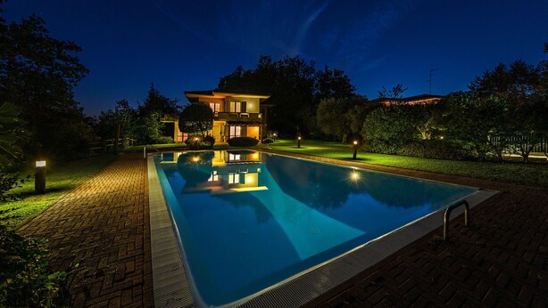 Una piscina con un camino de ladrillo que conduce a una casa de dos pisos con un balcón, rodeada de árboles y un cielo nocturno despejado.