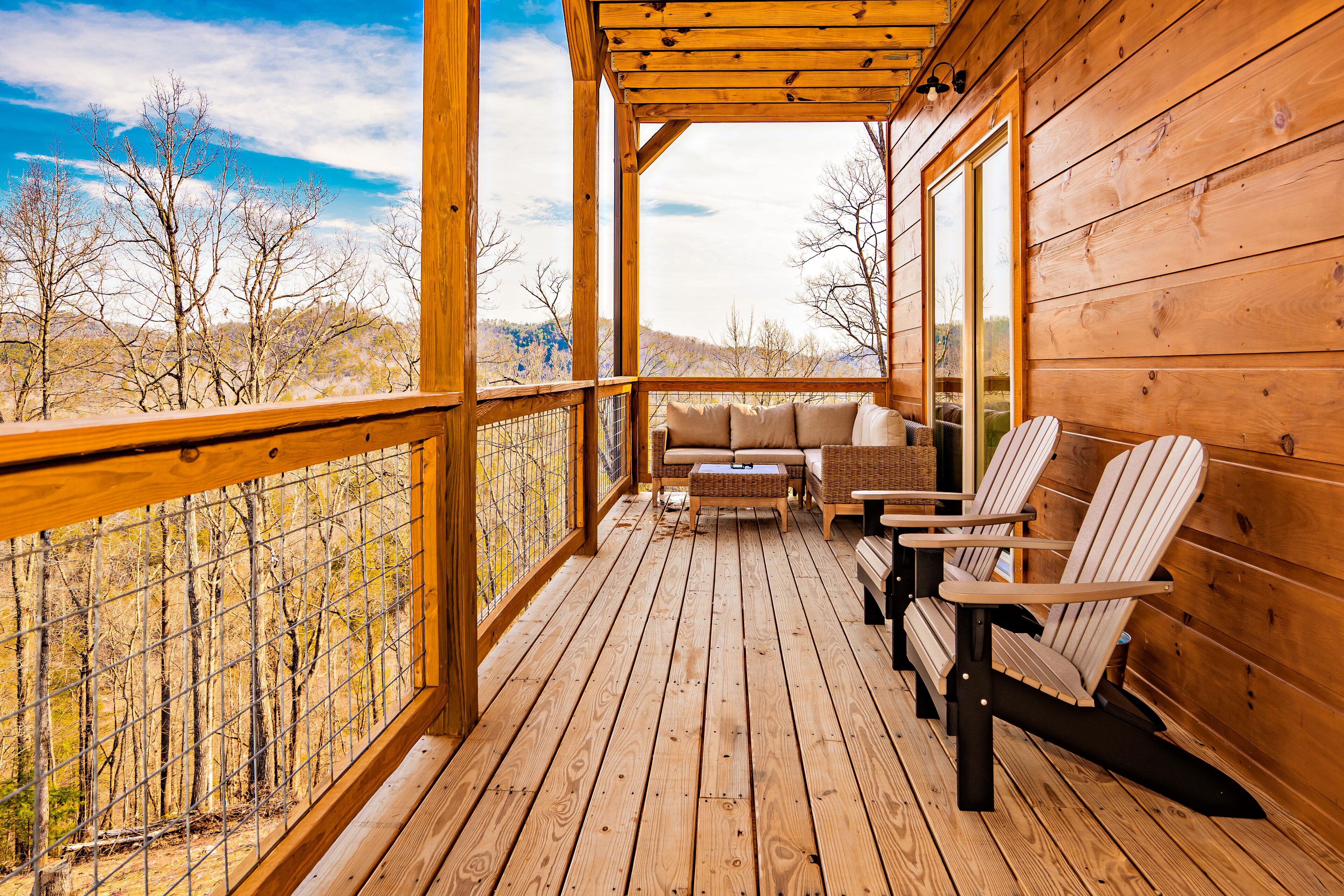 A wooden deck with a sofa and two Adirondack chairs, overlooking a forested area.