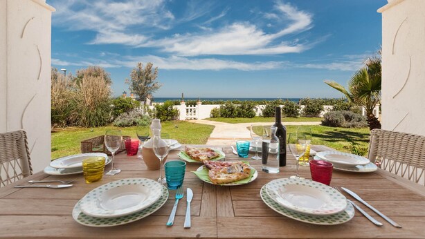 An outdoor dining table with plates, cutlery, and glasses, overlooking a garden and the sea.