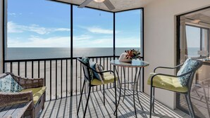 Un balcon avec vue sur la mer, des meubles en osier, un ventilateur de plafond et une petite table sur laquelle est posé un vase.