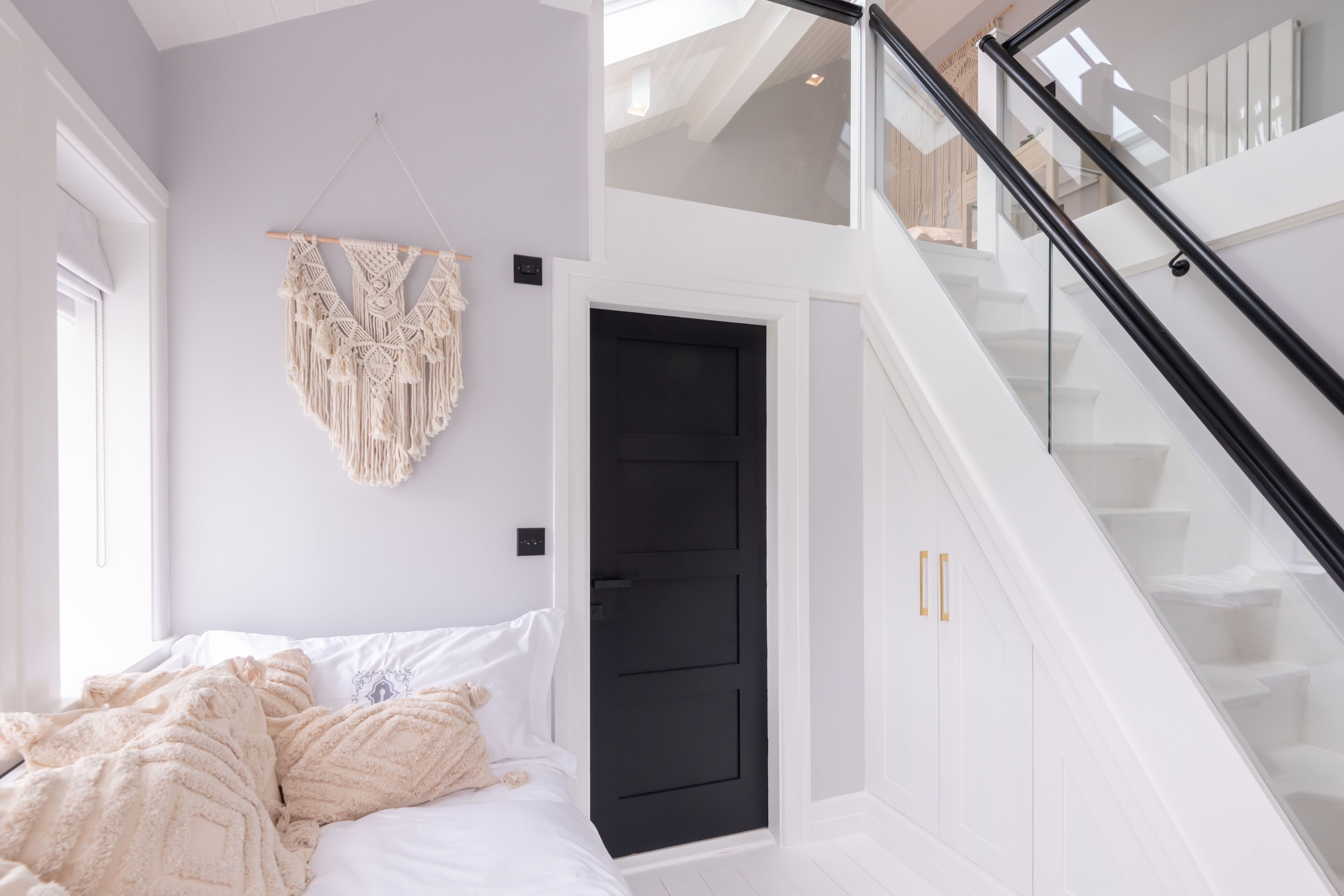 Une chambre avec un lit, une décoration murale en macramé, une porte noire et un escalier avec des rampes en verre.