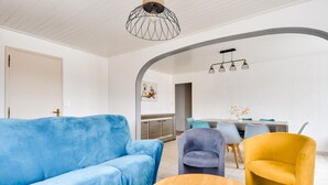 Un salon moderne avec un canapé bleu, un fauteuil jaune et une table basse en bois. On aperçoit à l’arrière-plan un coin repas avec une table et des chaises.