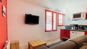 Un espacio habitable compacto con un sofá, una pequeña mesa de madera, un televisor, una kitchenette con microondas y una ventana con marcos rojos.