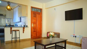 A living room with a wooden coffee table, a TV mounted on the wall, a wooden door, and a kitchen area with cabinets and a stove. - Mukoma Heights (Nairobi)