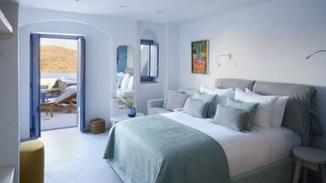 A bedroom with a large bed, a small table, a chair, and a view of the beach through an open door.