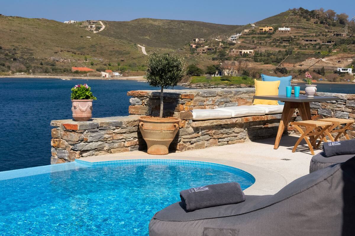 A private pool with stone walls, a lounge area with cushions, and a view of the sea and hills.