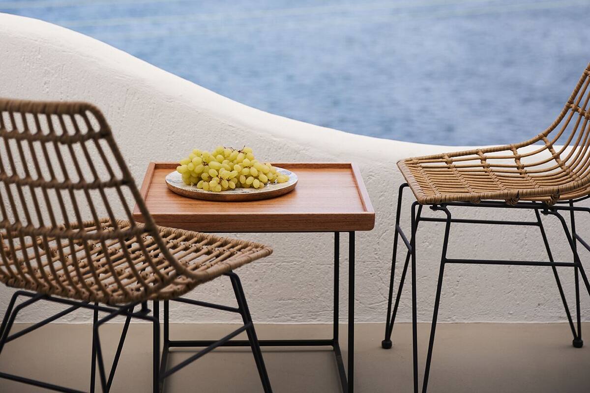 A terrace with two wicker chairs, a small wooden table, and a plate of grapes. The terrace overlooks a body of water and a distant hill with a building.