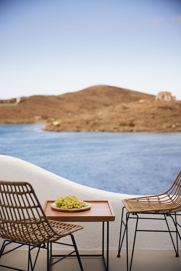 A terrace with two wicker chairs, a small wooden table, and a plate of grapes. The terrace overlooks a body of water and a distant hill with a building. - KEA MARE LUXURY VILLAS (Kea)