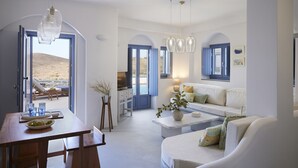 A modern living room with a sofa, coffee table, and dining area. There are blue doors leading to a balcony with a view of the landscape. - KEA MARE LUXURY VILLAS (Kea)