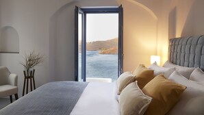 A bedroom with a large bed, a chair, a lamp, and a view of the sea through an open door. - KEA MARE LUXURY VILLAS (Kea)