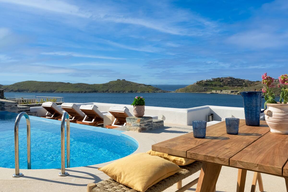 A poolside area with lounge chairs, a wooden table, and a view of the sea and hills.