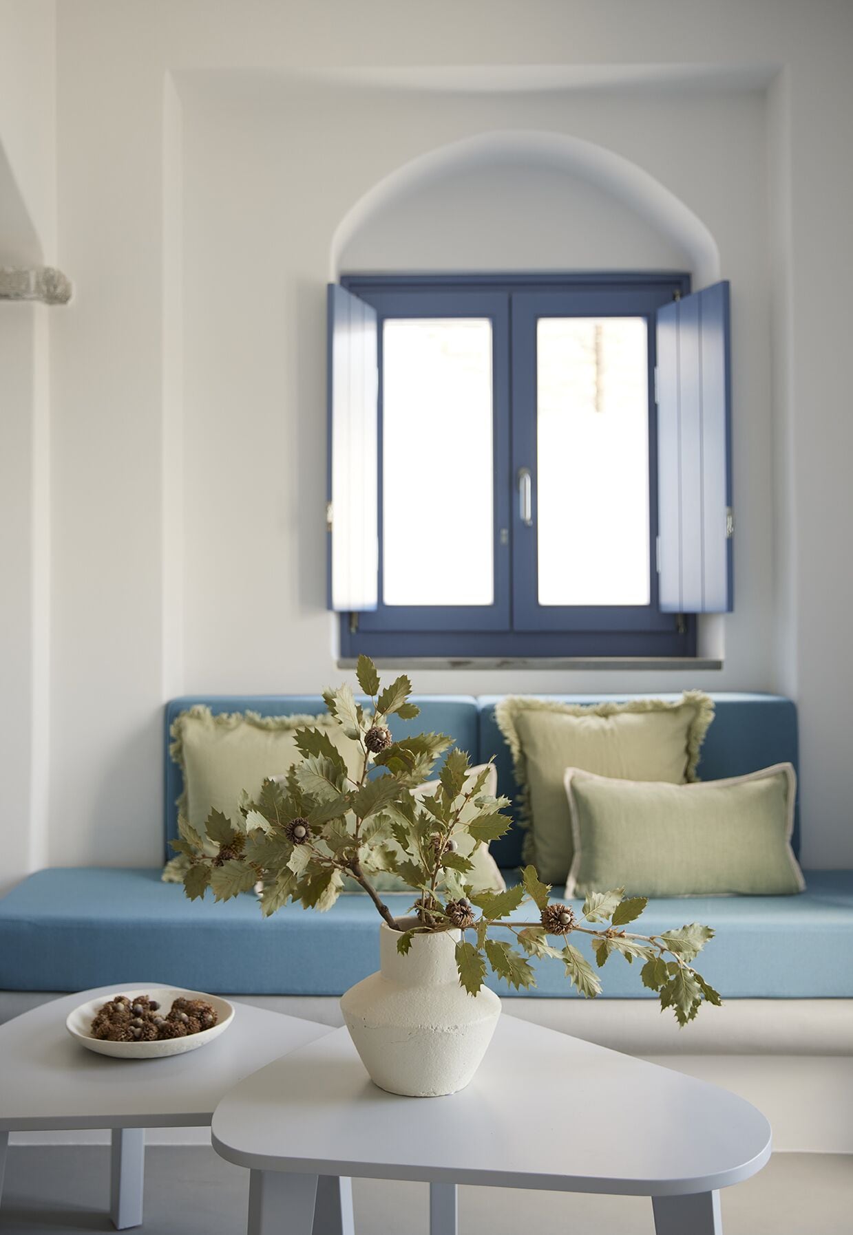 A room with a white table, a white vase with green leaves, a blue sofa with light green cushions, and a window with blue shutters.