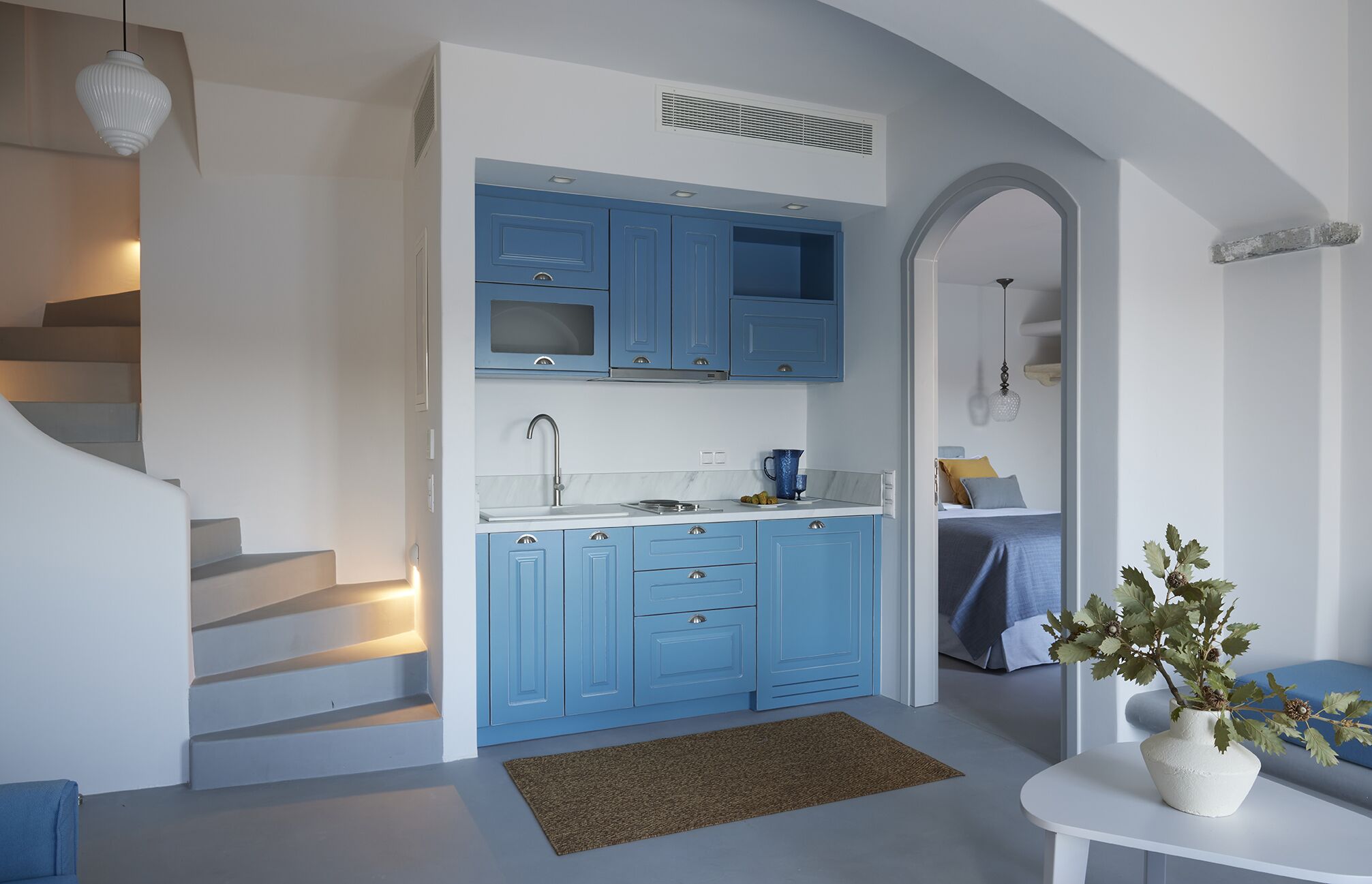 A modern kitchen with blue cabinets, a white countertop, and a built-in oven. There is a staircase leading to another level and a small table with a vase of flowers.