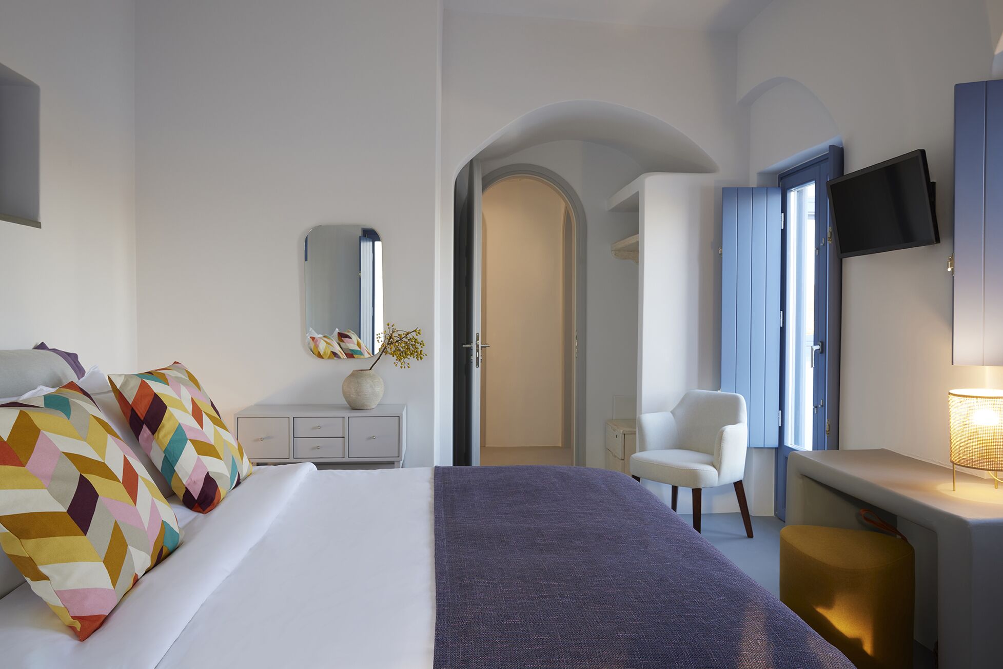 A modern hotel room with a bed, a desk, a chair, a TV, and a door leading to another room.