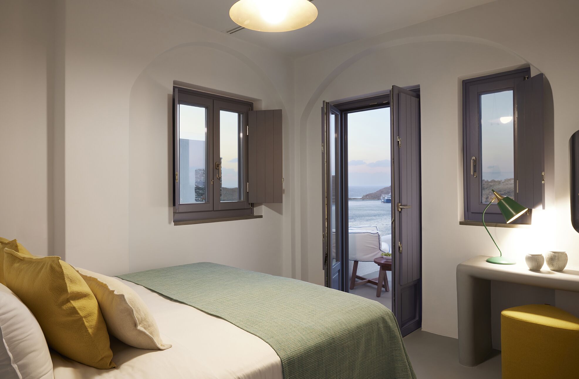 A bedroom with a bed, a desk, and a view of the sea through open doors.