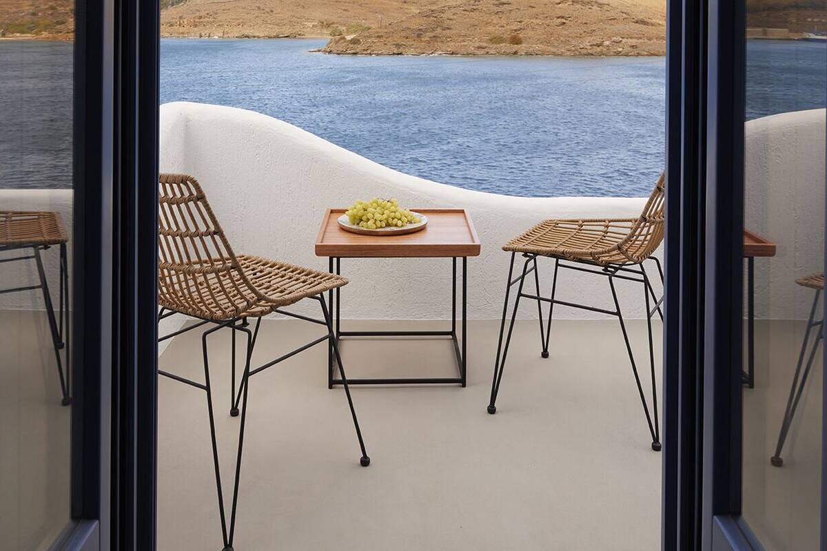 A balcony with wicker chairs and a table, overlooking a body of water and a hilly landscape.