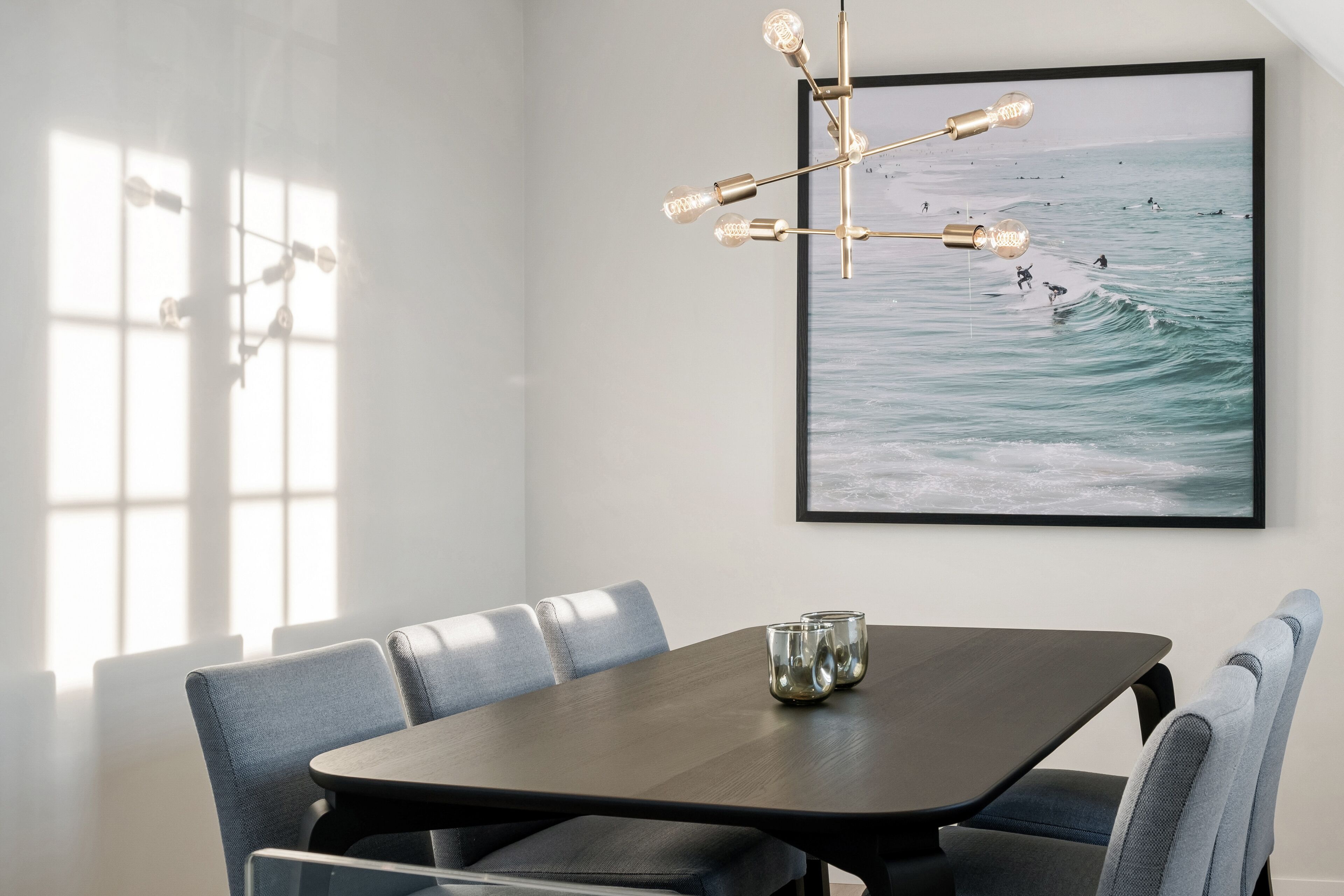 Un comedor moderno con una mesa de madera oscura, sillas tapizadas en gris y un cuadro con vista al océano.