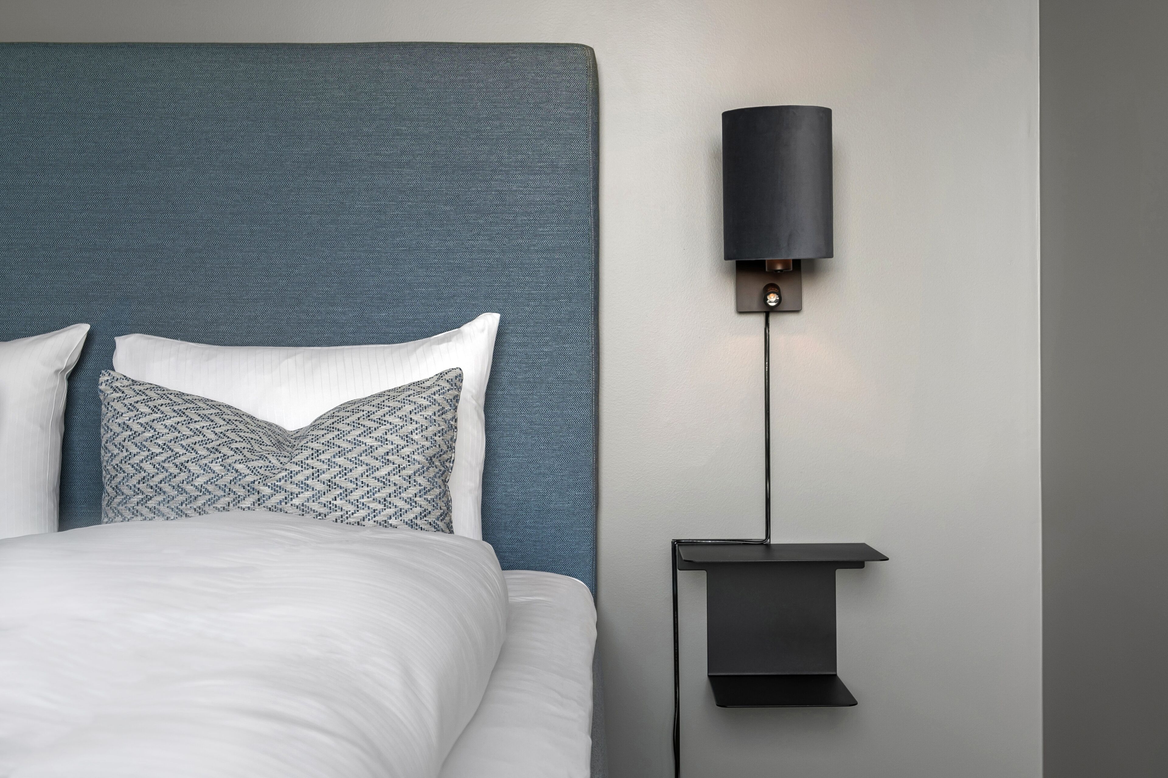 Uma cama bem arrumada com cabeceira azul, almofada estampada e luminária fixada na parede.
