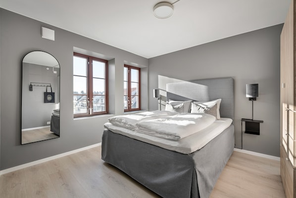 A modern bedroom with a large bed, a wall-mounted mirror, and two windows.