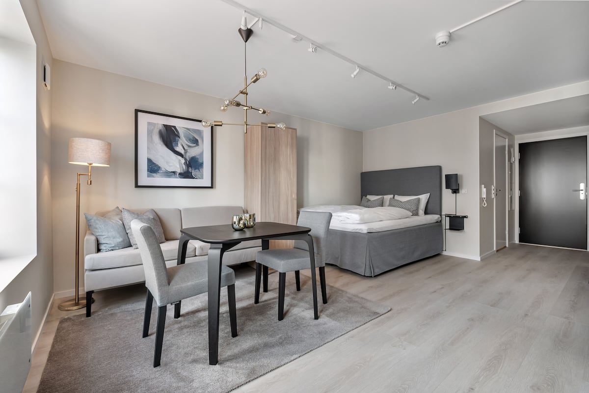 A modern hotel room with a sofa, a dining table, a bed, and a large window.