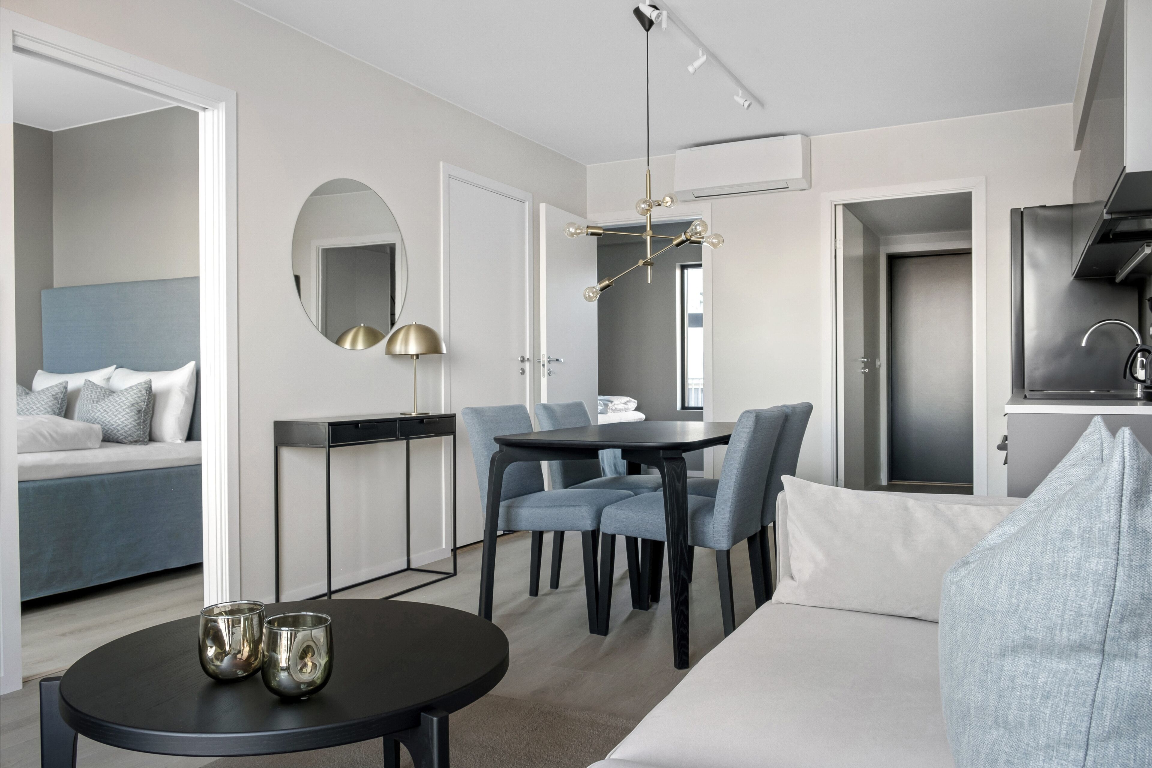 A modern hotel room with a bed, a dining table with chairs, a small round table with two glasses, a mirror, and a ceiling light fixture.