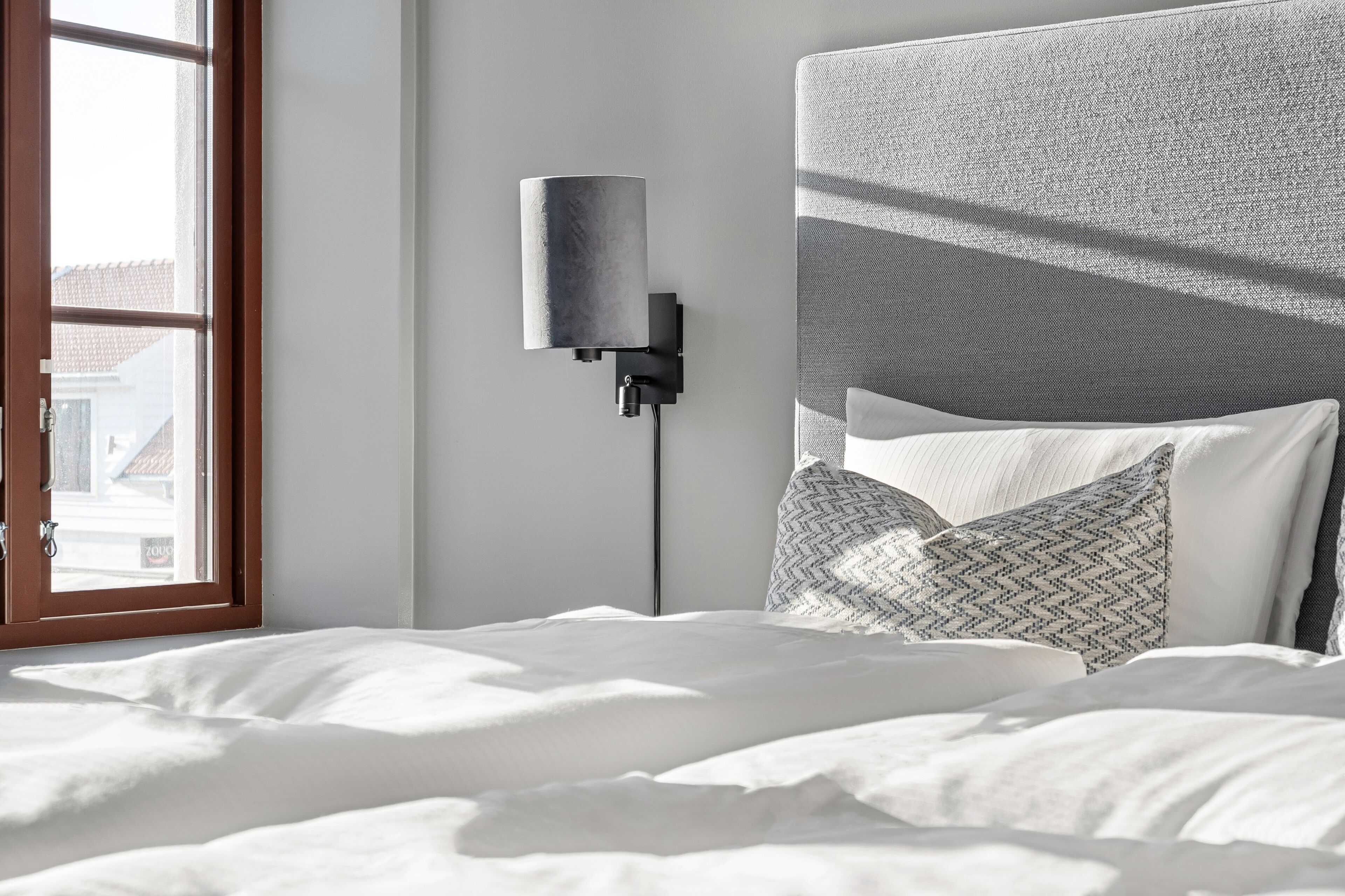 Una cama bien hecha con sábanas blancas, un cabecero gris con textura y almohadas estampadas. Se ve una lámpara moderna fijada a la pared a la izquierda.