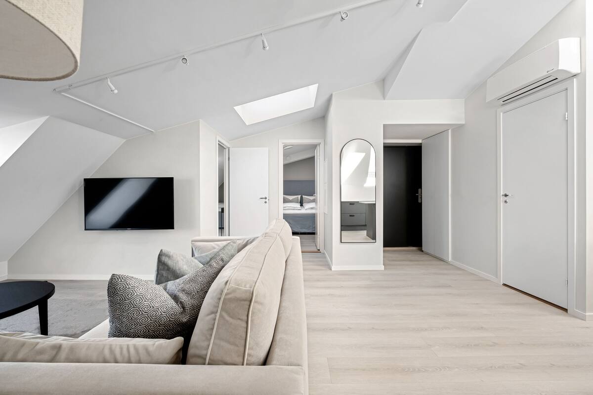 A modern loft apartment with a high ceiling, skylight, a flat-screen TV, a sofa with patterned cushions, and a minimalist design.