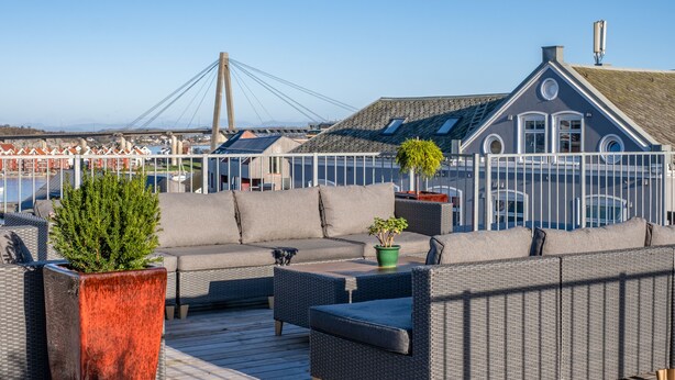 A deck with outdoor furniture, a potted plant, and a view of a bridge and buildings in the background. - VANDER - Pedersgata (Stavanger)